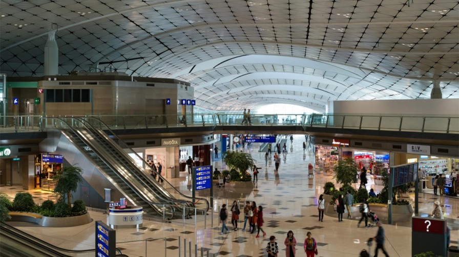 A spacious and modern airport terminal with passengers walking around, shops, digital signage, and escalators, connecting Zhuhai and Hong Kong airports via a cross-border coach service.
